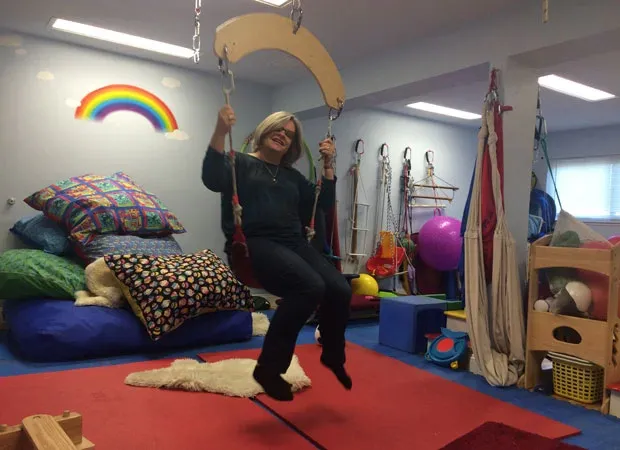 Janet Carney on a platform swing in the therapy room, a rainbow painted on the wall behind her